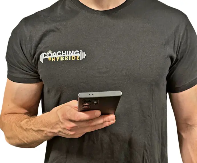 alt="Coach sportif tenant un smartphone avec l'application Coaching Hybride, vêtu d'un t-shirt à l'effigie de sa marque"